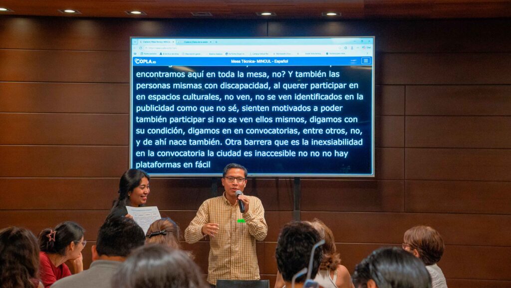 Un hombre sostiene un micrófono y habla frente a un grupo de personas durante una mesa de trabajo sobre accesibilidad. A su lado, una mujer sostiene una hoja mientras lo acompaña. Detrás de ellos hay una pantalla grande que muestra subtítulos en tiempo real con texto blanco sobre fondo negro. El público permanece sentado observando la intervención.