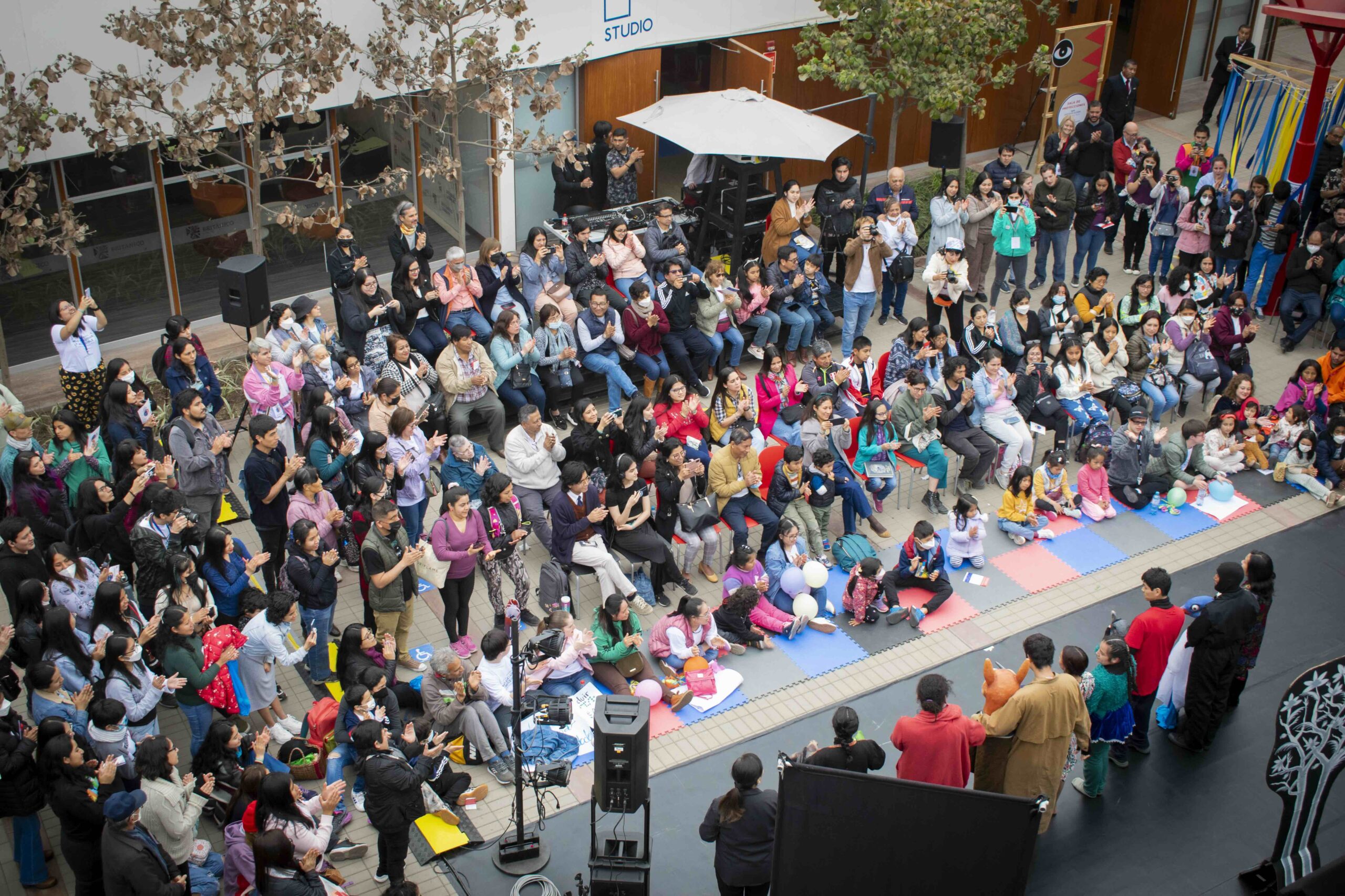 Fotografía panorámica de un patio interior lleno de personas reunidas. El público, compuesto por personas de distintas edades, aplaude mientras observa una actividad o presentación en un escenario ubicado en la parte inferior derecha. En el centro hay varias filas de sillas y, en el suelo, infancias sentadas sobre colchonetas de colores con globos. Se observan equipos de sonido, cámaras y elementos escenográficos alrededor del espacio.