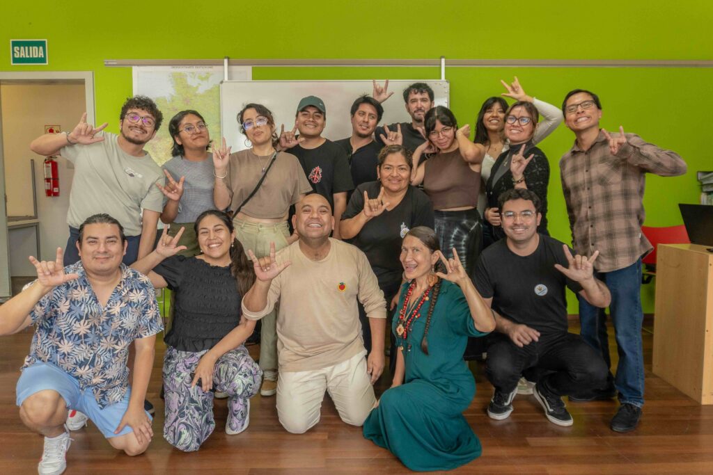 Fotografía grupal de 16 personas posando sonrientes en un aula con paredes verdes, frente a una pizarra blanca. Varias personas están de pie en la parte posterior y otras se encuentran arrodilladas o agachadas en la parte delantera. La mayoría realiza señas con las manos, incluyendo el gesto de «te amo» en Lengua de Señas Peruana. Visten ropa casual de distintos colores. Al fondo se observan un mapa, una puerta con el letrero «SALIDA» y parte del mobiliario del aula. La imagen transmite un ambiente de compañerismo y celebración.