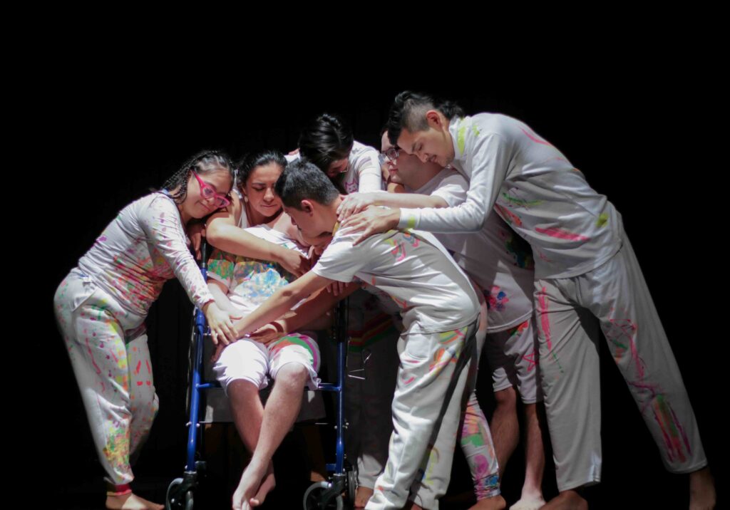 Seis personas en escena, vestidas de blanco con manchas y trazos de colores, se agrupan alrededor de una persona sentada en una silla de ruedas. Varias personas extienden sus brazos y manos hacia ella, creando una composición colectiva de apoyo y cercanía. La escena está iluminada sobre un fondo negro y transmite una sensación de encuentro, cuidado y comunidad.