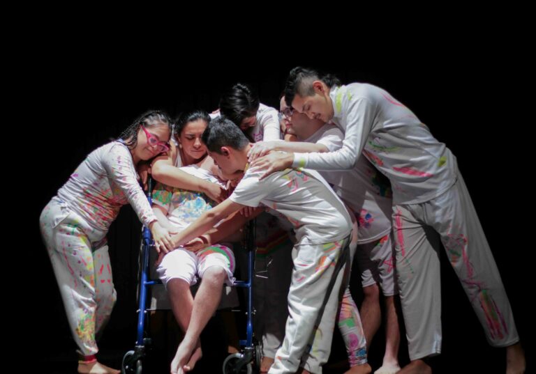 Seis personas en escena, vestidas de blanco con manchas y trazos de colores, se agrupan alrededor de una persona sentada en una silla de ruedas. Varias personas extienden sus brazos y manos hacia ella, creando una composición colectiva de apoyo y cercanía. La escena está iluminada sobre un fondo negro y transmite una sensación de encuentro, cuidado y comunidad.