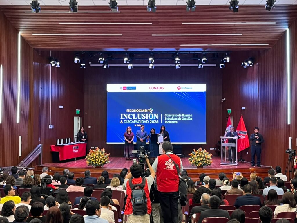 La directora de CAPAZ Perú, Déborah Grandez Seminario, recibe un diploma de reconocimiento en el escenario durante la ceremonia del «Reconocimiento Inclusión & Discapacidad 2026» de CONADIS, frente a un auditorio lleno de gente.
